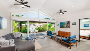 Un salon spacieux au design moderne, comprenant un canapé gris, des fauteuils bleus, un meuble en bois et une porte en verre donnant sur un patio aménagé avec du mobilier d’extérieur.