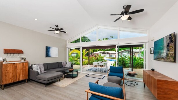 Un vaste salon doté d’un canapé gris, de fauteuils bleus et d’un meuble en bois. De grandes fenêtres offrent une vue sur un jardin luxuriant et une piscine.