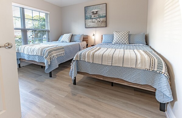 A bedroom with two beds, wooden flooring, a window with blinds, and a painting on the wall.