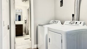 A laundry room with two front-loading washing machines and a dryer, a door leading to a bathroom, and a vent on the wall.