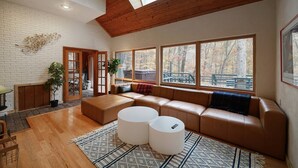 Ein modernes Wohnzimmer mit einem Ecksofa, zwei runden weißen Hocker und großen Fenstern mit Blick auf Bäume.