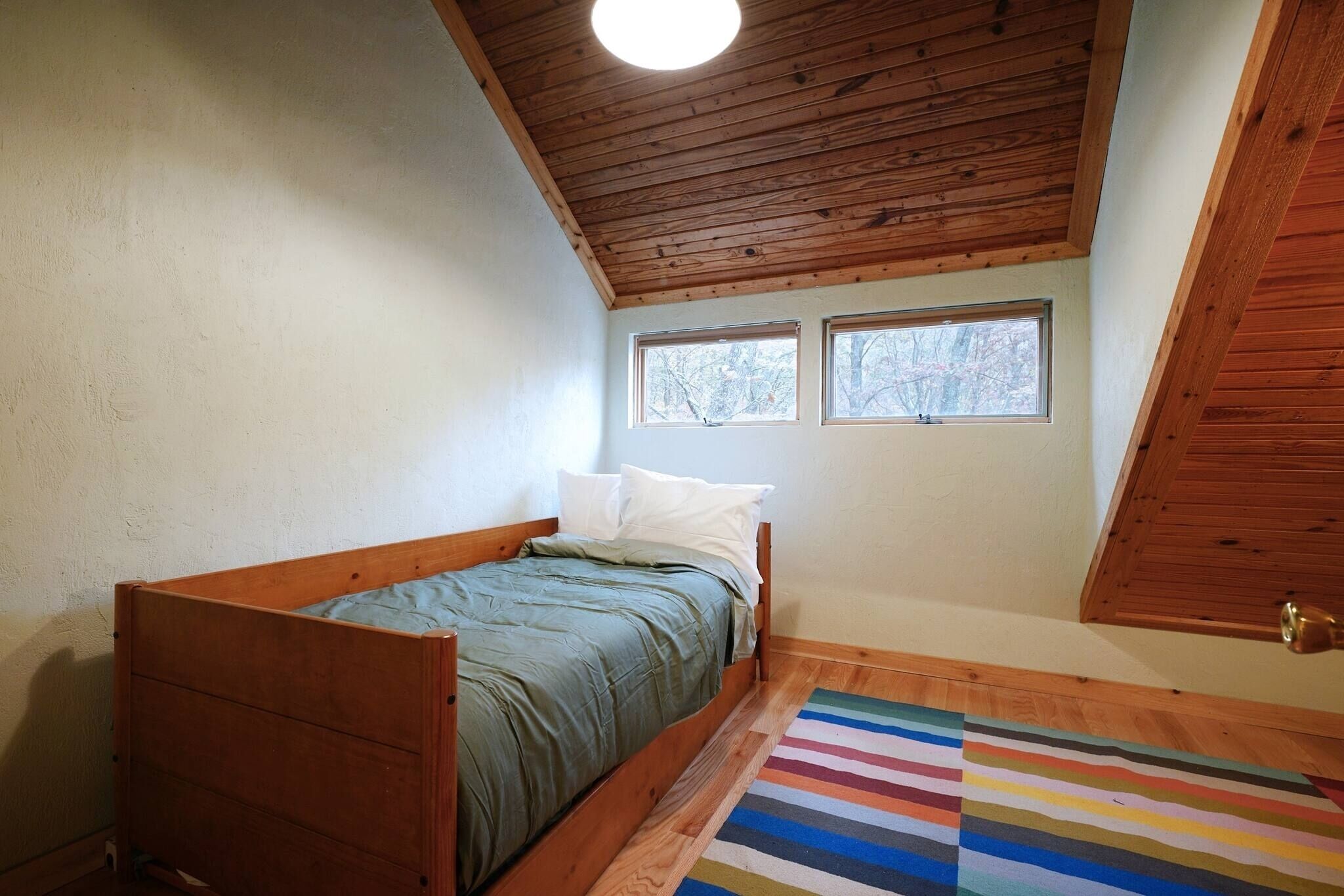 Une petite chambre avec un lit simple, un plafond en bois, deux fenêtres et un tapis rayé aux couleurs vives.