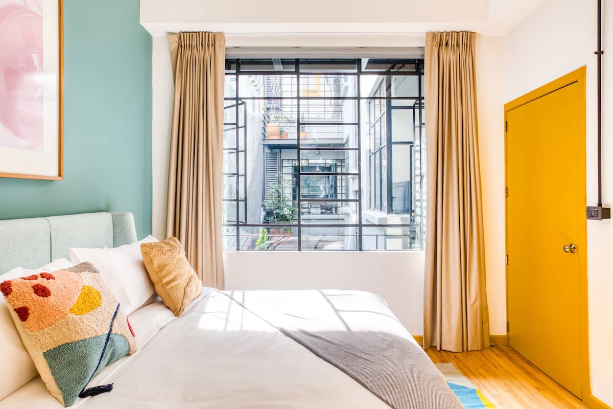 Hotellihuone, jossa on sänky, ikkuna verhoilla, keltainen ovi ja näkymä kaupungin ylle.
