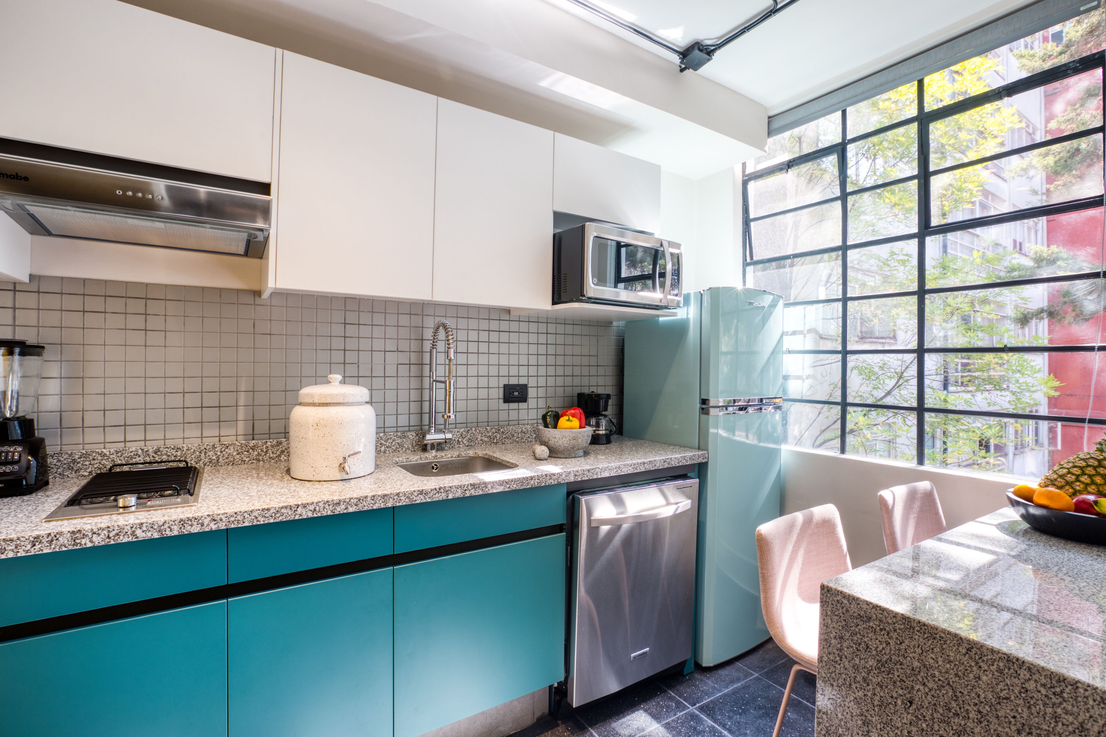 Una cocina moderna con armarios color turquesa, un lavavajillas de acero inoxidable, un microondas y un amplio ventanal con vistas a la ciudad.