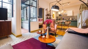 Una sala de estar moderna con un sofá, una mesa de centro y un sillón con un jarrón de flores. Hay un televisor sobre un mueble de madera y se distingue una zona de cocina al fondo.