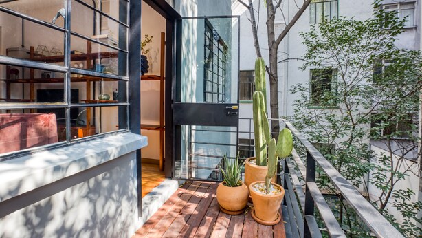 Un balcón con piso de madera, dos cactus en macetas y un ventanal de vidrio que da a una habitación con interior de madera.