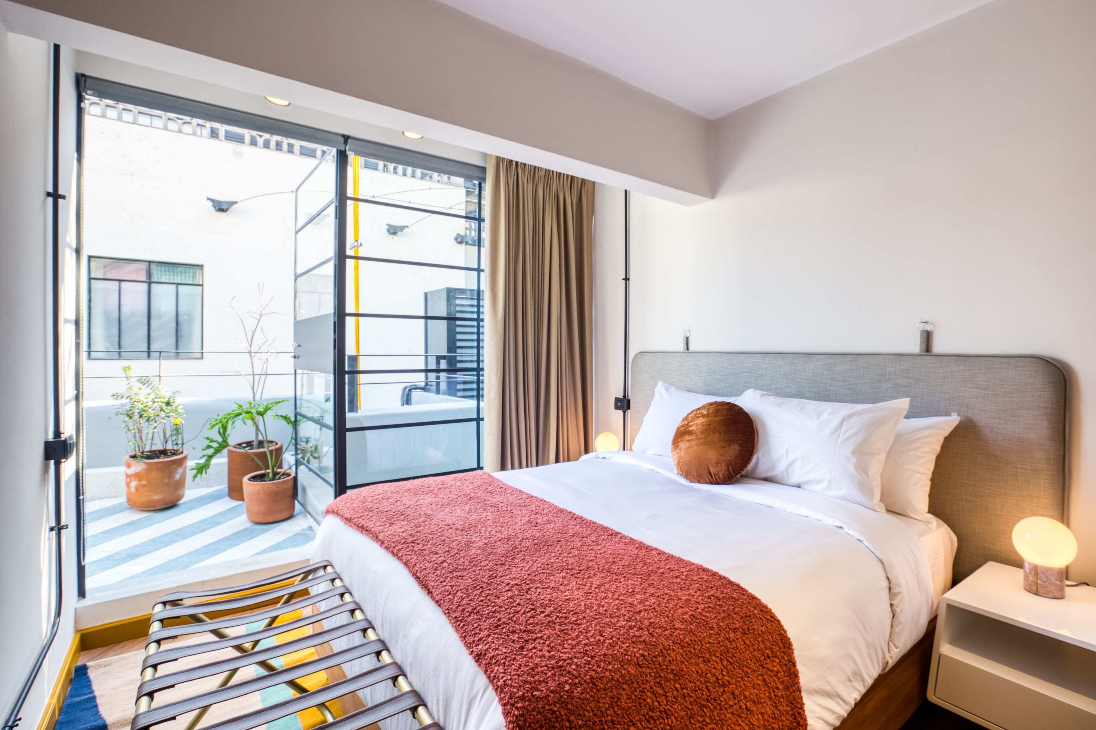 Habitación de hotel con cama, mesita de noche, lámpara, ventana con cortinas y vistas al exterior.