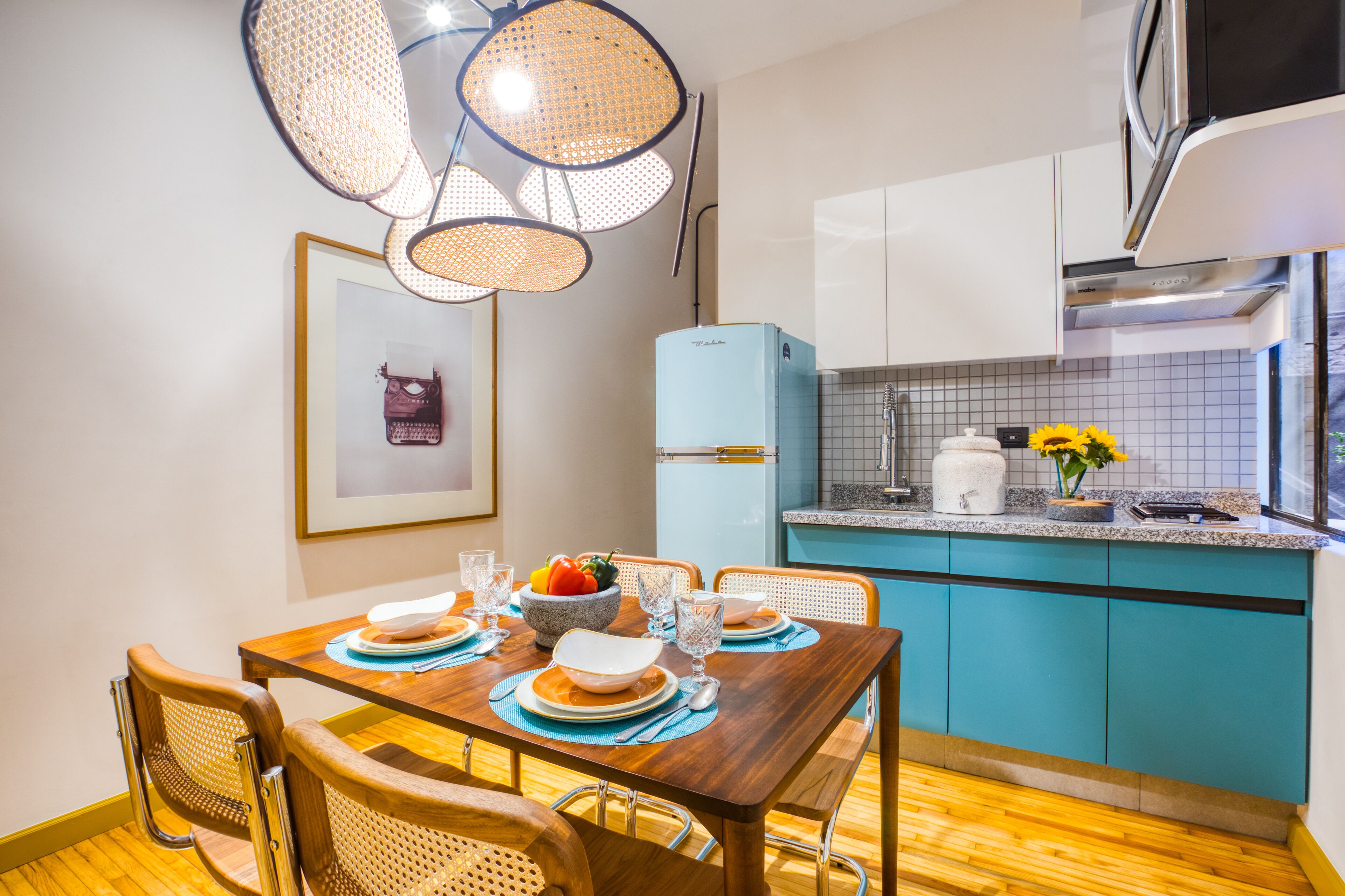 Un coin repas avec une table en bois dressée pour quatre personnes, un réfrigérateur bleu et une cuisine avec des armoires de couleur turquoise et un plan de travail blanc.