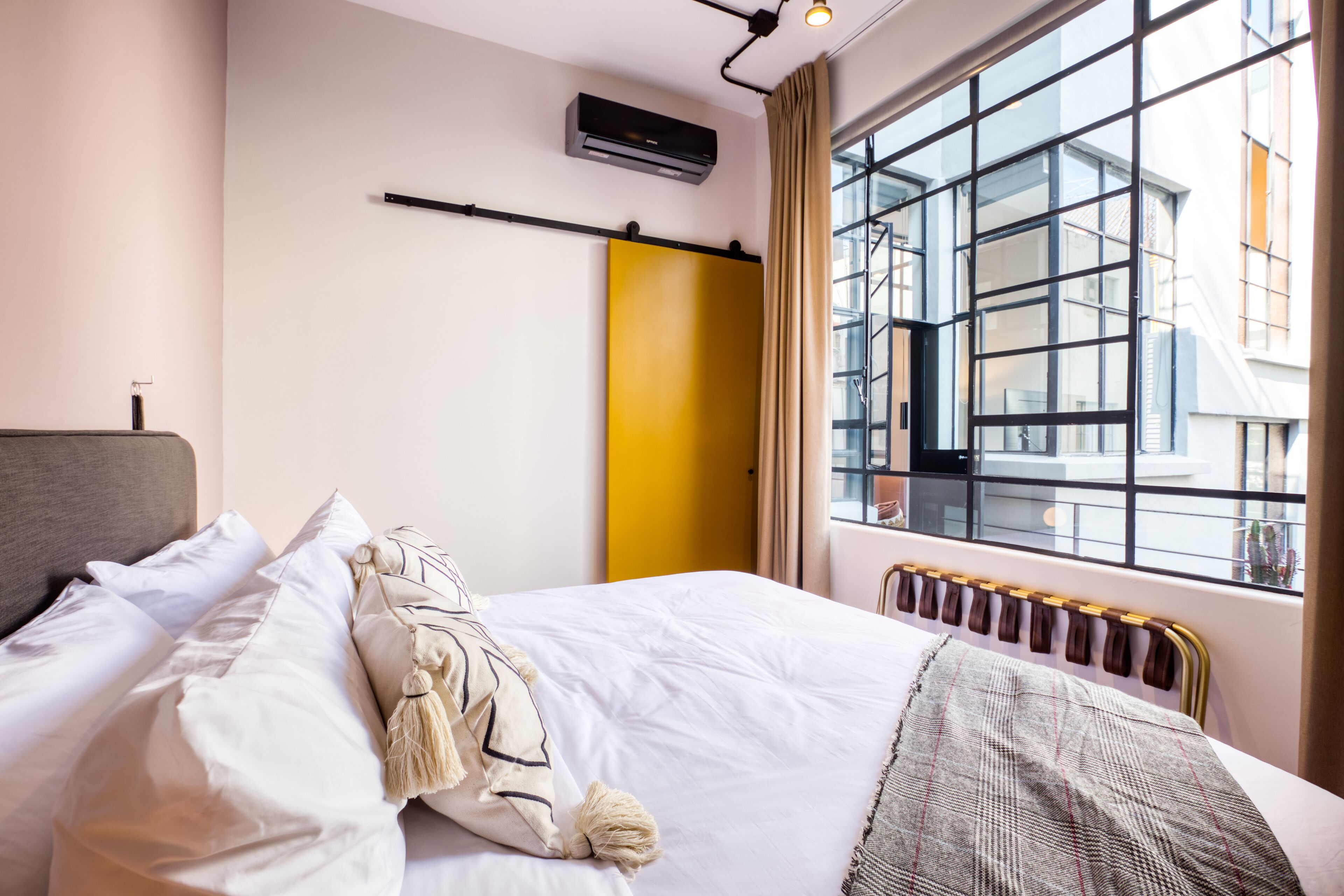 Hotellihuone, jossa on sänky, patteri, ikkuna, josta on näkymä rakennuksiin, ja seinälle asennettu ilmastointilaite.