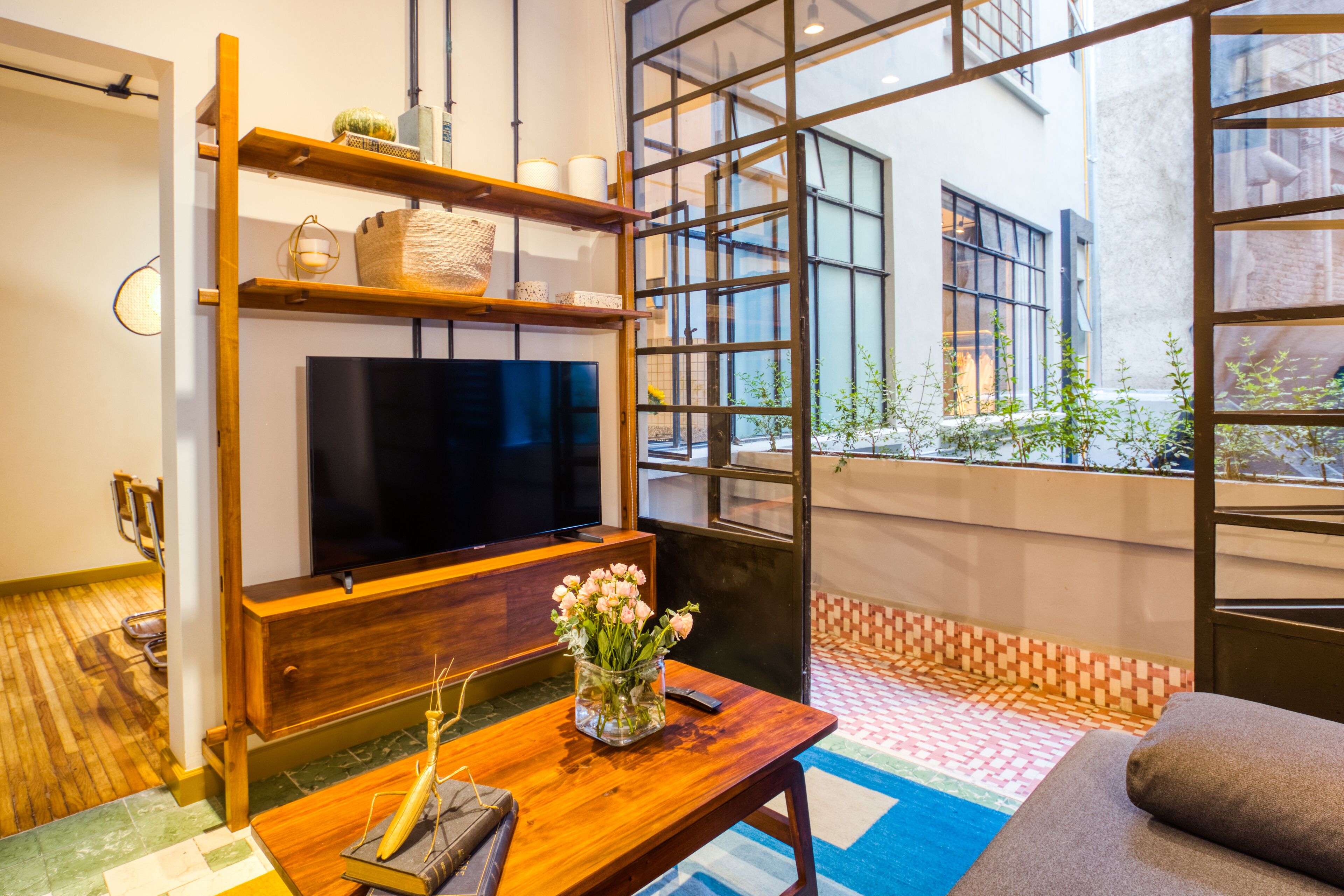 A modern living room with a flat-screen TV, wooden shelves, a coffee table with a vase of flowers, and a view of the city through large windows.
