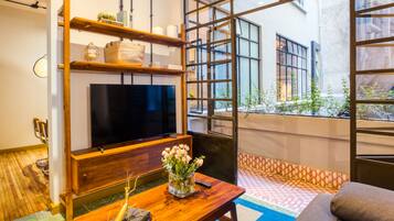 A modern living room with a flat-screen TV, wooden shelves, a coffee table with a vase of flowers, and a view of the city through large windows.