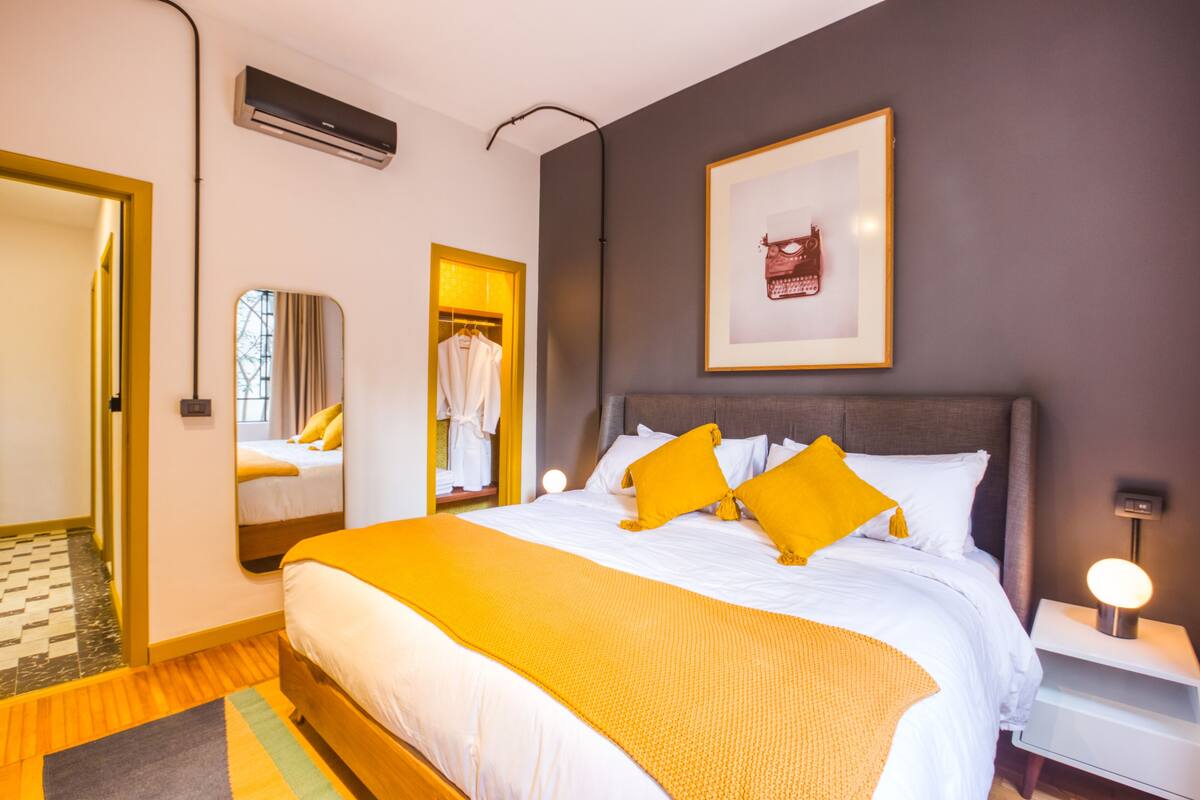 Hotellihuone, jossa on suuri sänky, yöpöytä lampulla, vaatekaappi, peili ja ilmastointilaite.