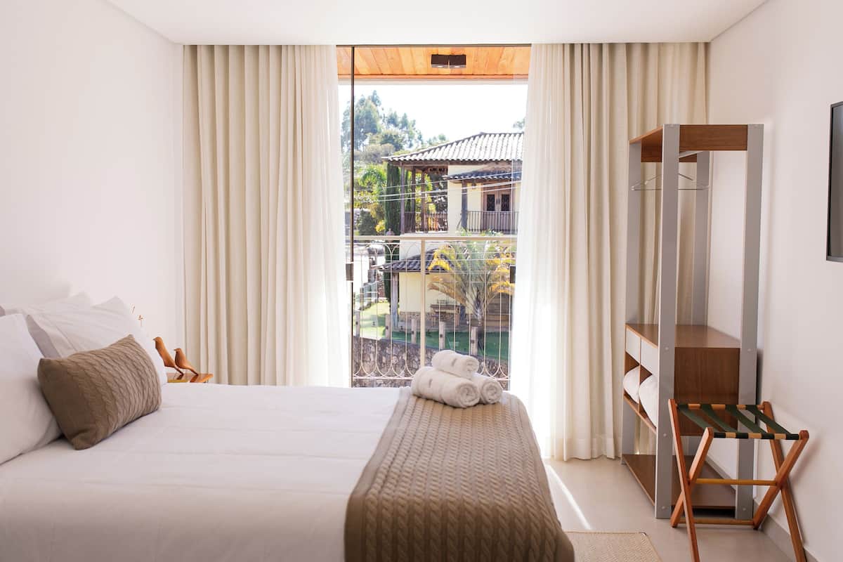 A hotel room with a large bed, a wooden bench, and a view of a building through the window.