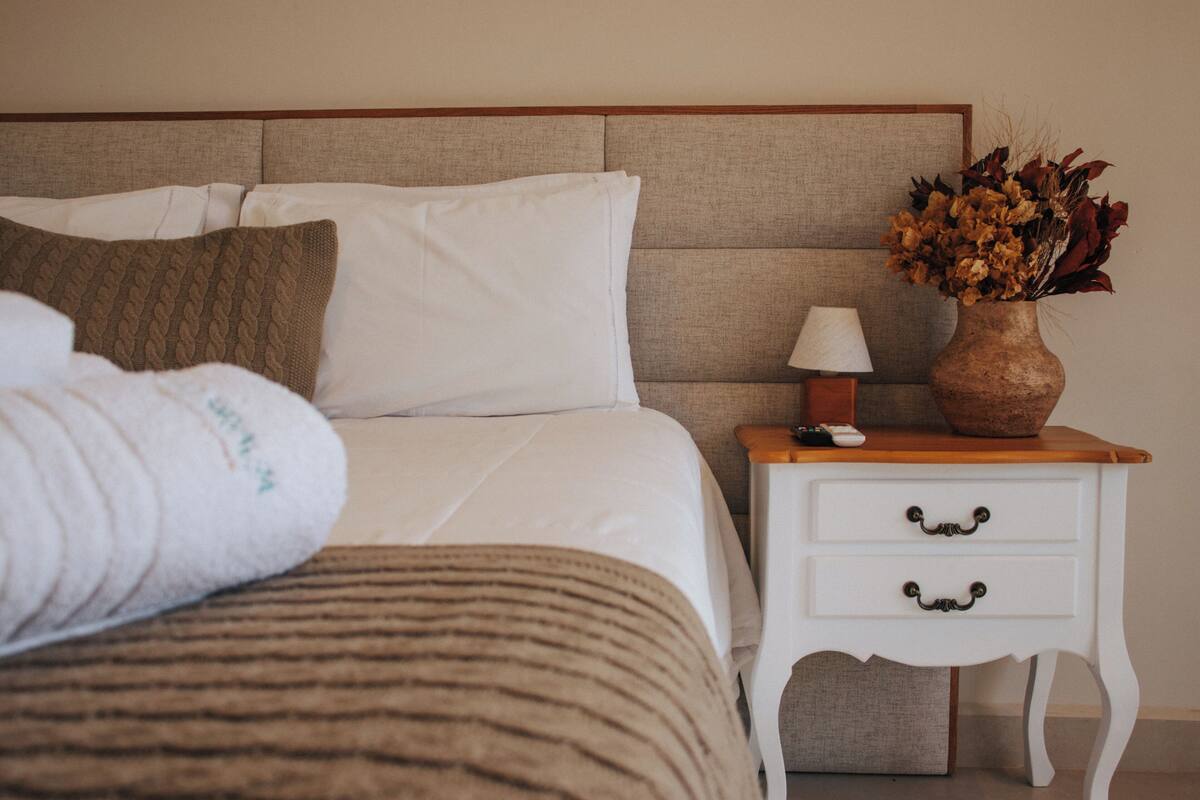 A neatly made bed with white and brown bedding, a bedside table with a lamp, and a vase with dried flowers.