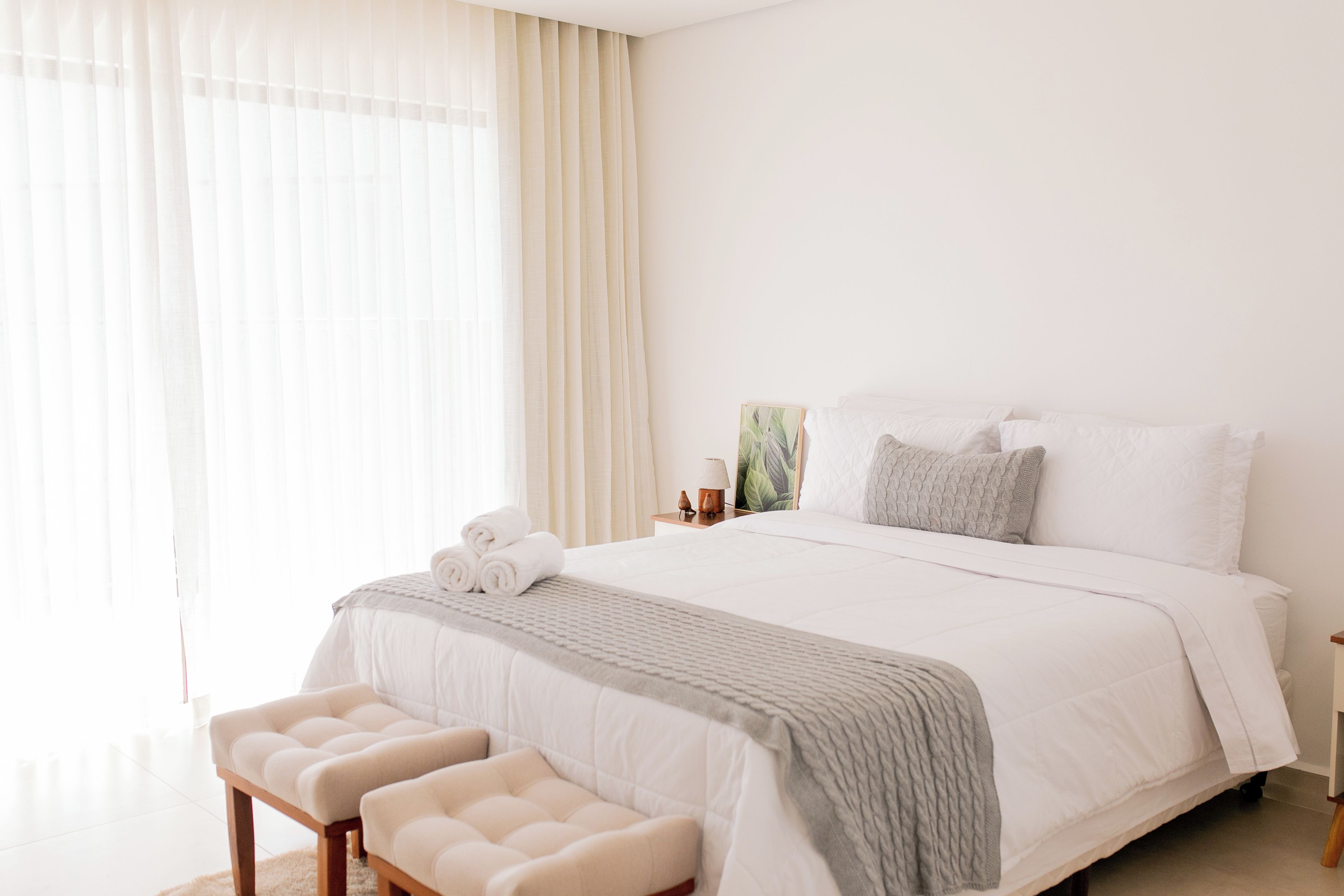 A hotel room with a large bed, a bedside table with a lamp, a small plant, and a window with sheer curtains.
