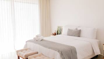 A hotel room with a large bed, a bedside table with a lamp, a small plant, and a window with sheer curtains.