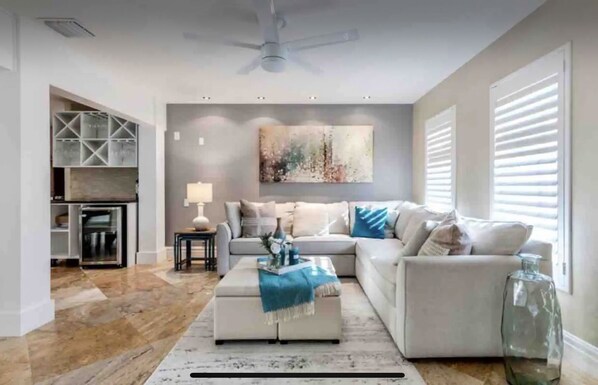 A modern living room with a sofa, ottoman, and a glass coffee table. There is a painting on the wall and a lamp on a side table.