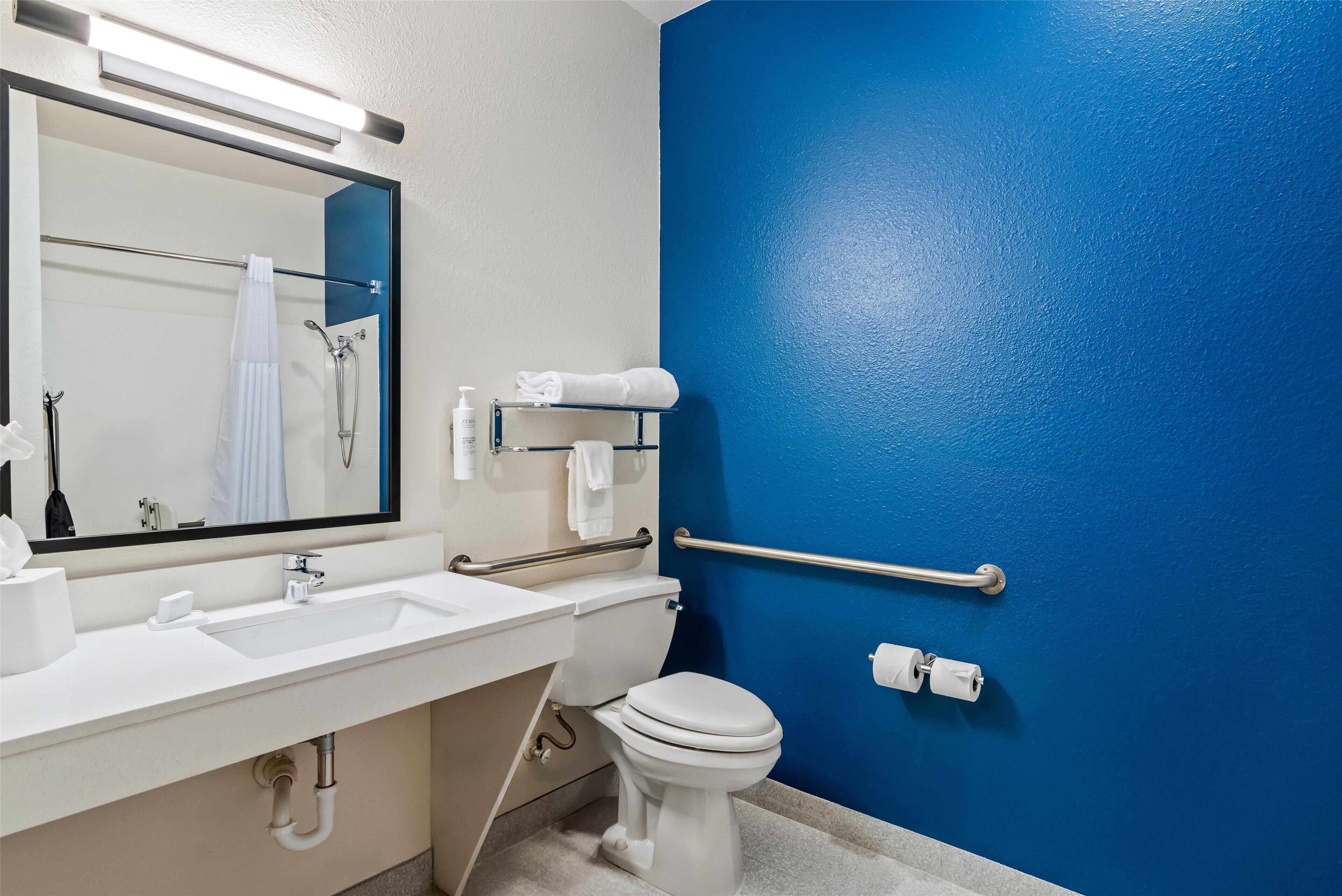 Une salle de bain avec un lavabo blanc, un miroir et des toilettes, un mur bleu et une barre d’appui.