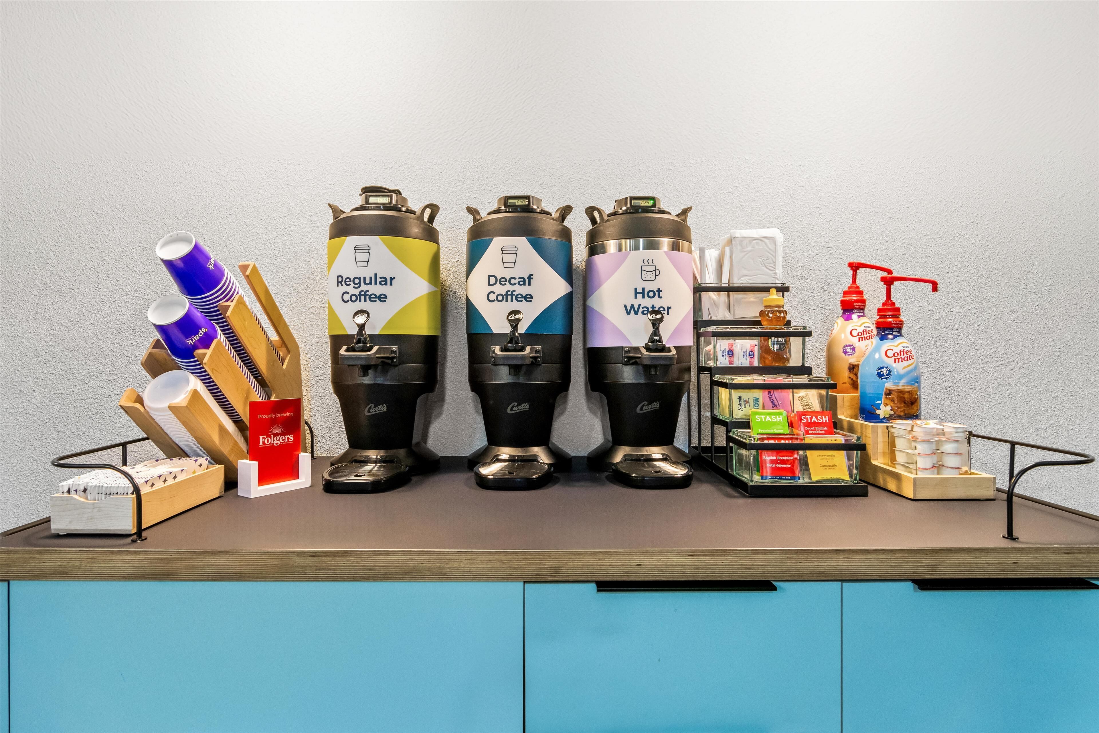 A coffee station with dispensers for regular coffee, decaf coffee, and hot water, along with coffee mugs, napkins, and a soap dispenser.