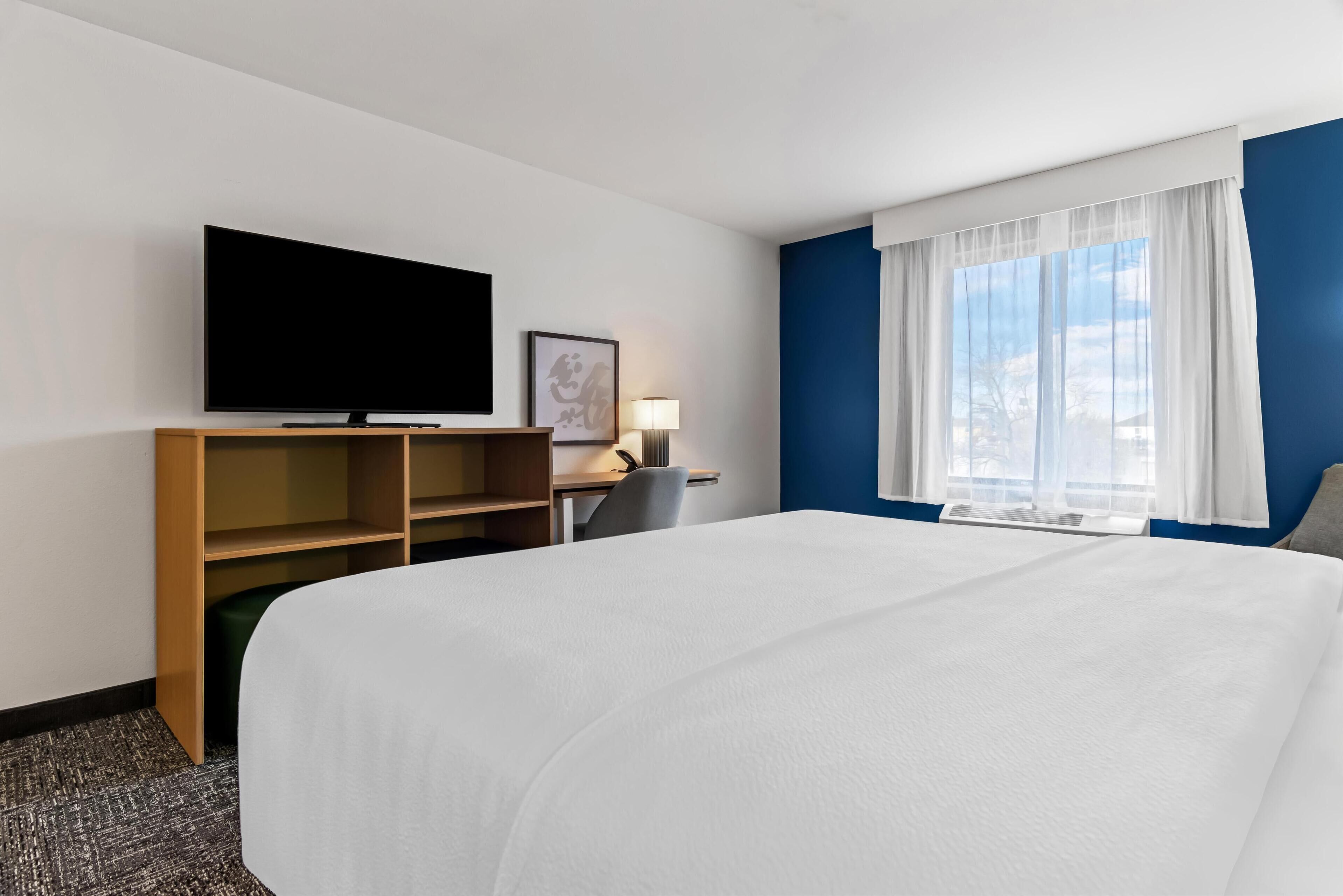 A hotel room with a large bed, a flat-screen TV, a desk with a chair, and a window with curtains.