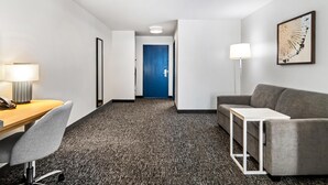 Una habitación de hotel con escritorio, silla, sofá gris y una mesita con lámpara.