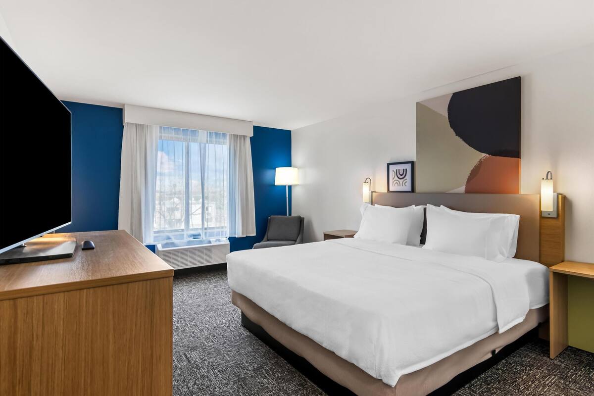 A hotel room with a large bed, a desk with a computer, a chair, a window with curtains, and a painting on the wall.