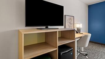 Habitación de hotel con televisor de pantalla plana montado en un estante de madera, un escritorio con lámpara, una silla y un refrigerador pequeño.