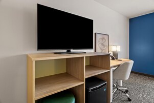 Habitación de hotel con televisor de pantalla plana montado en un estante de madera, un escritorio con lámpara, una silla y un refrigerador pequeño.