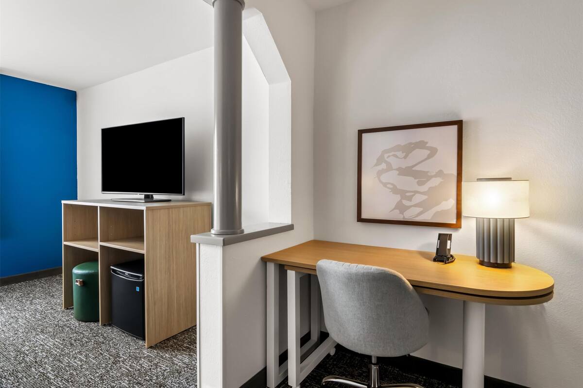 A hotel room with a TV, a desk with a lamp, a chair, and a trash bin.