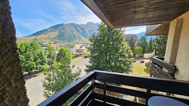 Un balcón con vistas a un paisaje montañoso, un área pavimentada y un edificio con techo inclinado.