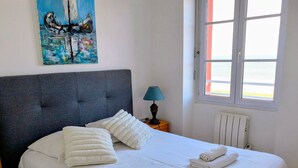 Ein ordentlich bezogenes Bett mit weißen Leinen und Kissen, ein Gemälde an der Wand, eine Nachttischlampe und ein Fenster mit Aussicht.
