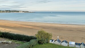 Ein Strand mit einer Reihe kleiner, weißer Strandhütten mit blauen Dächern, einem ruhigen Meer und einer entfernten Küstenlinie mit Gebäuden.