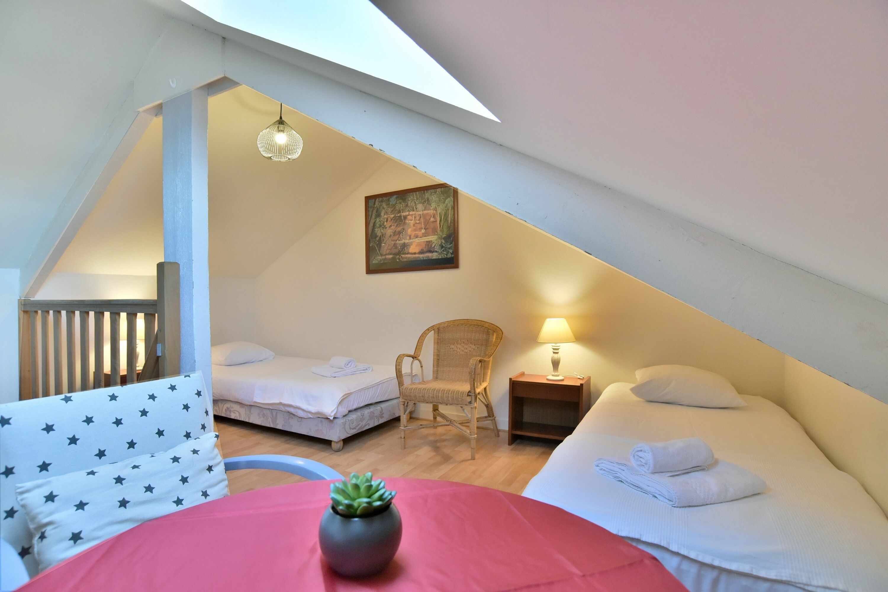 Une chambre mansardée accueillante, avec deux lits, un fauteuil en osier, une petite table avec une lampe et une plante en pot.