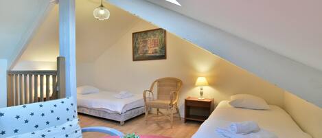 Une chambre mansardée accueillante, avec deux lits, un fauteuil en osier, une petite table avec une lampe et une plante en pot.