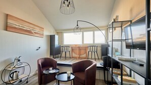 A modern living room with a wooden floor, two leather armchairs, a small round table, a coffee table, a bookshelf, and a television.