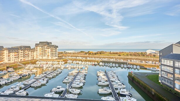 A marina with numerous boats docked in calm water, surrounded by buildings and a clear sky.