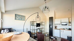 A modern kitchen with white cabinets, a microwave, and a coffee machine. There is a dining area with a wooden table and chairs.