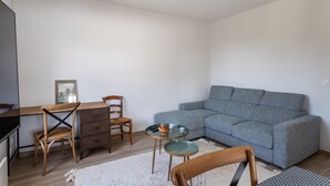 Een woonkamer met een grijze bank, houten stoelen, een kleine ronde tafel met een mand en een beker, en een bureau met een stoel.