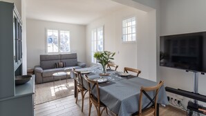 Een moderne woonkamer met een eethoek, een grijze bank, een flatscreen-tv en grote ramen.