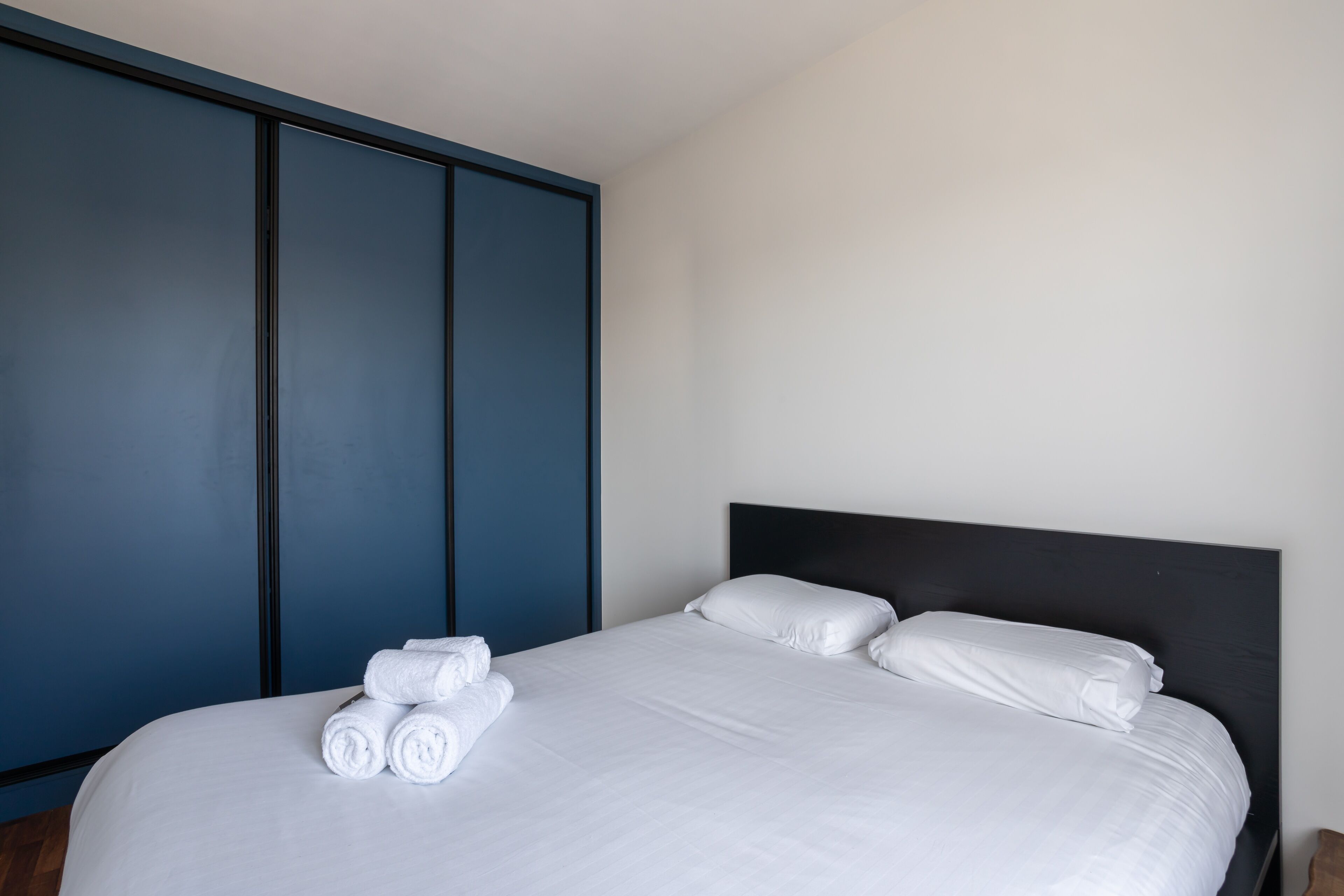 Una cama bien hecha con sábanas y almohadas blancas, un cabecero oscuro y un armario azul al fondo.