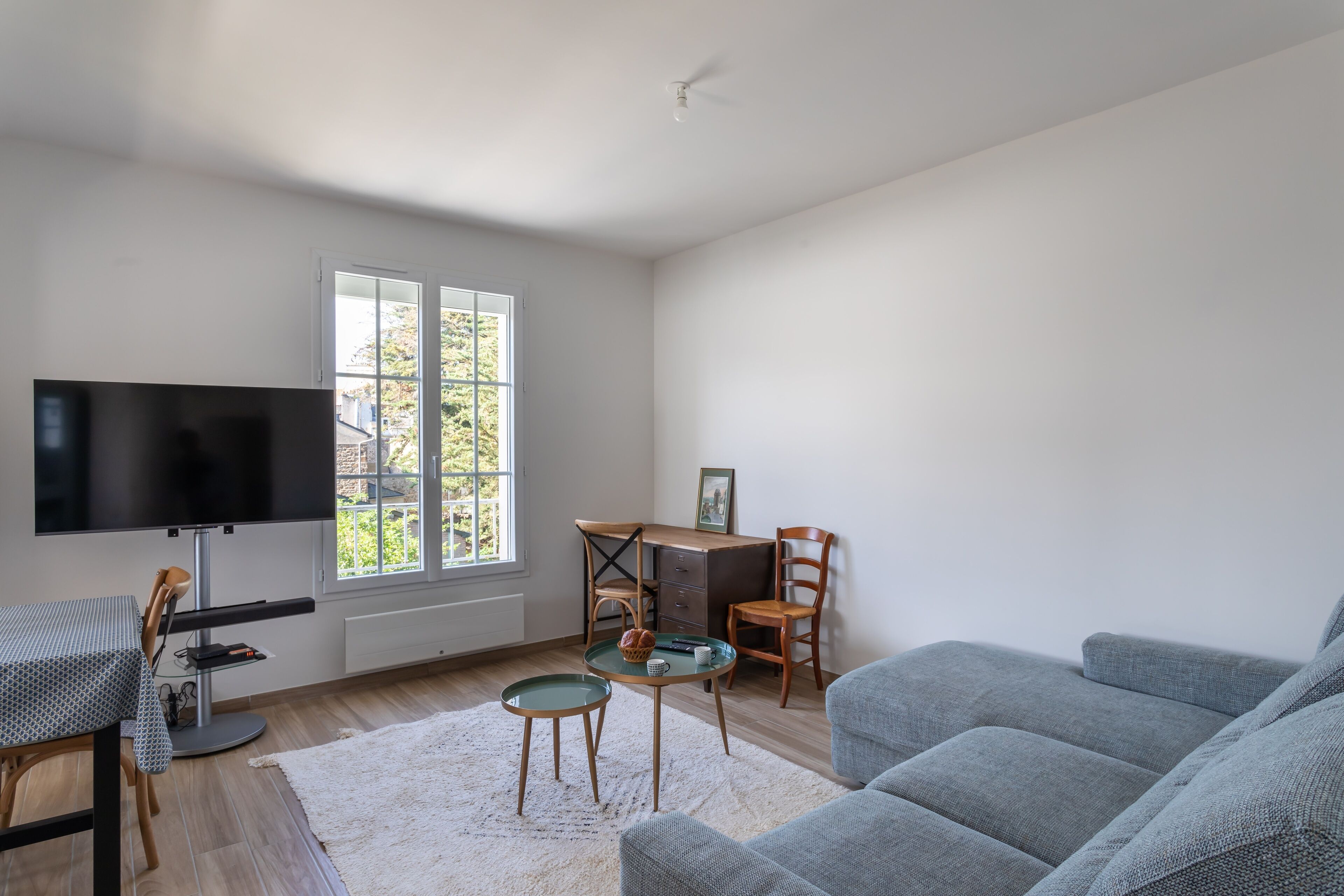 A living room with a TV, a desk, two chairs, a sofa, and a window with a view.