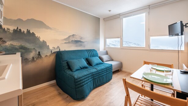 Une pièce chaleureuse avec un canapé de couleur turquoise, une table à manger avec des chaises et une fresque représentant un paysage de montagne sur le mur.