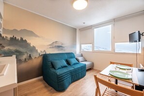 A cozy room with a teal sofa, a dining table with chairs, and a mural of a mountain landscape on the wall.