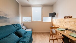 Une pièce chaleureuse avec un canapé de couleur turquoise, une table à manger en bois et des chaises, un téléviseur fixé au mur et une fenêtre donnant sur les montagnes.