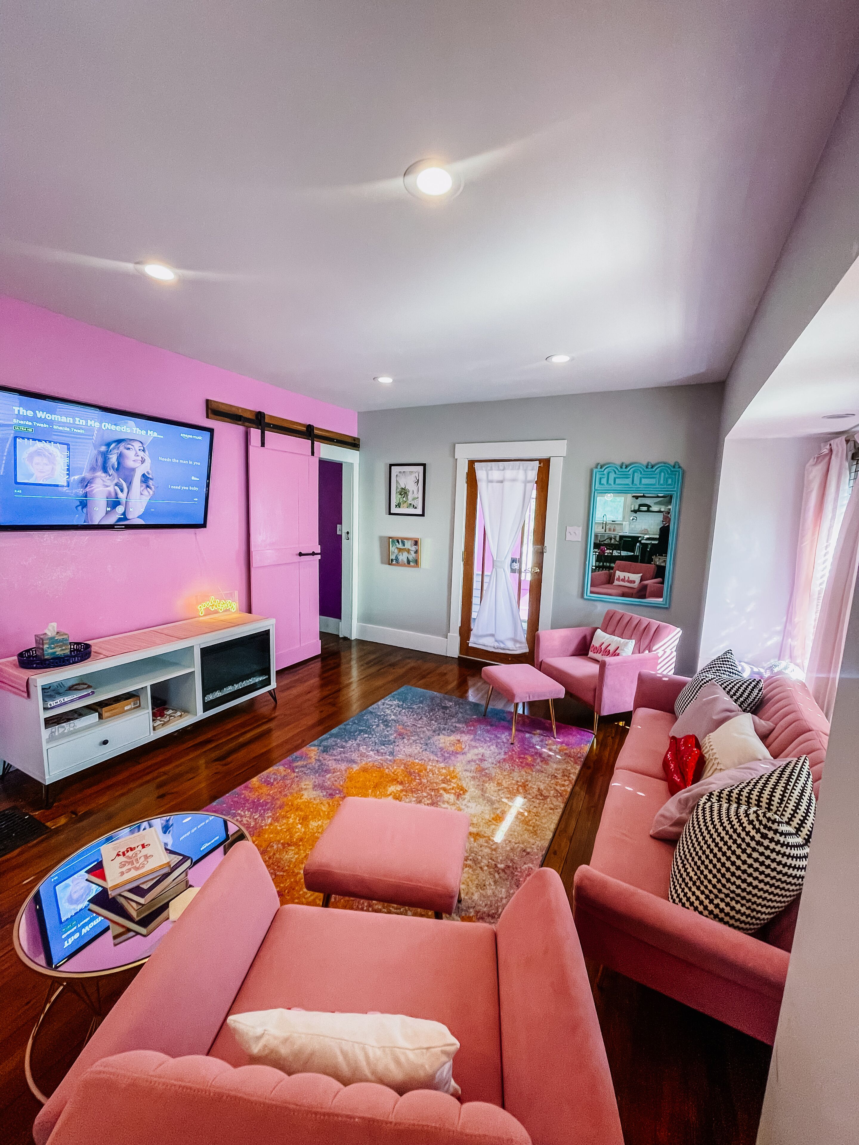 A living room with a pink sofa, a TV on the wall, a wooden floor, and a colorful rug.