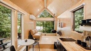 A compact, modern cabin with a wooden interior, a cozy seating area, a small kitchen, and large windows offering a view of the forest.