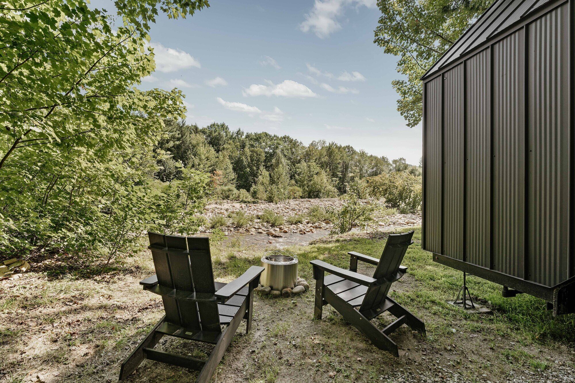 Un espace extérieur aménagé dans un style rustique, avec deux fauteuils Adirondack et un foyer en métal, offrant une vue sur une rivière et une zone boisée.