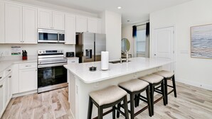 A modern kitchen with white cabinets, stainless steel appliances, a marble island with bar stools, and a view to the living area. - Lovely Villa near Disney | Margaritaville & Waterpark Access - 2956OR (Kissimmee)