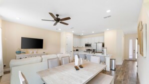 Un salon moderne doté d’un téléviseur à écran plat, d’un ventilateur de plafond, d’une table à manger et d’une cuisine en arrière-plan.