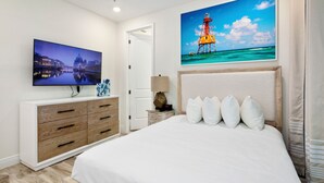 Een slaapkamer met een grote flatscreen televisie, een houten dressoir, een nachtkastje met een lamp en een bed met witte beddengoed en kussens.